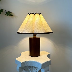 K&ouml;nnte beinhalten: Eine braune Tischlampe mit einem wei&szlig;en Leinenlampenschirm und einem schwarzen Besatz. Die Lampe steht auf einem wei&szlig;en Sockel.