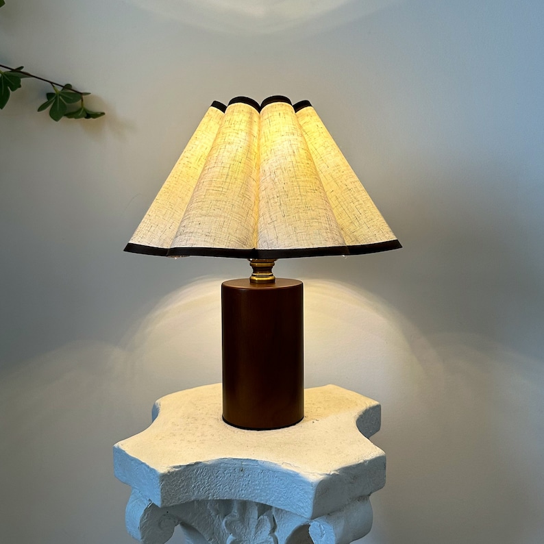K&ouml;nnte beinhalten: Eine braune Tischlampe mit einem wei&szlig;en Leinenlampenschirm und einem schwarzen Besatz. Die Lampe steht auf einem wei&szlig;en Sockel.