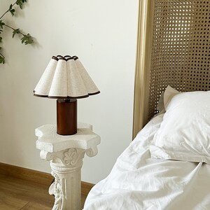 K&ouml;nnte beinhalten: Ein wei&szlig;er Lampenschirm mit braunem Besatz steht auf einem wei&szlig;en Podesttisch. Der Lampenschirm ist plissiert und hat einen braunen Holzfu&szlig;. Die Lampe steht vor einem wei&szlig;en Bett mit einem Korbgeflecht-Kopfteil.