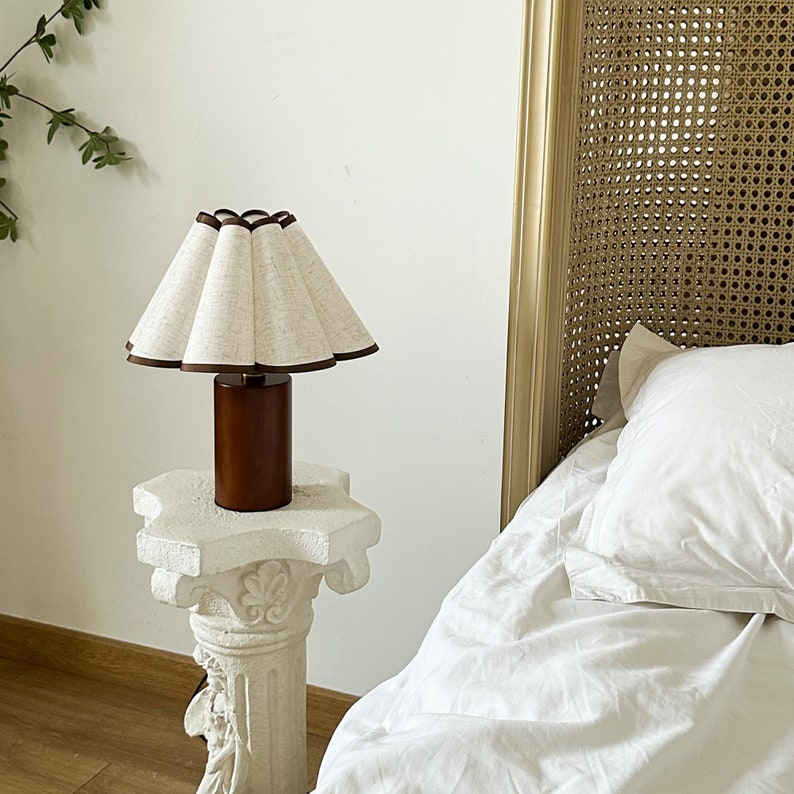 K&ouml;nnte beinhalten: Ein wei&szlig;er Lampenschirm mit braunem Besatz steht auf einem wei&szlig;en Podesttisch. Der Lampenschirm ist plissiert und hat einen braunen Holzfu&szlig;. Die Lampe steht vor einem wei&szlig;en Bett mit einem Korbgeflecht-Kopfteil.