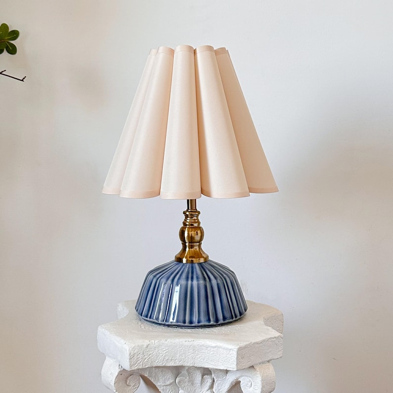 May include: A decorative table lamp with a pleated, light pink lampshade. The lamp features a brass base and a blue ceramic body with vertical ridges. The lamp is a stylish home decor item.