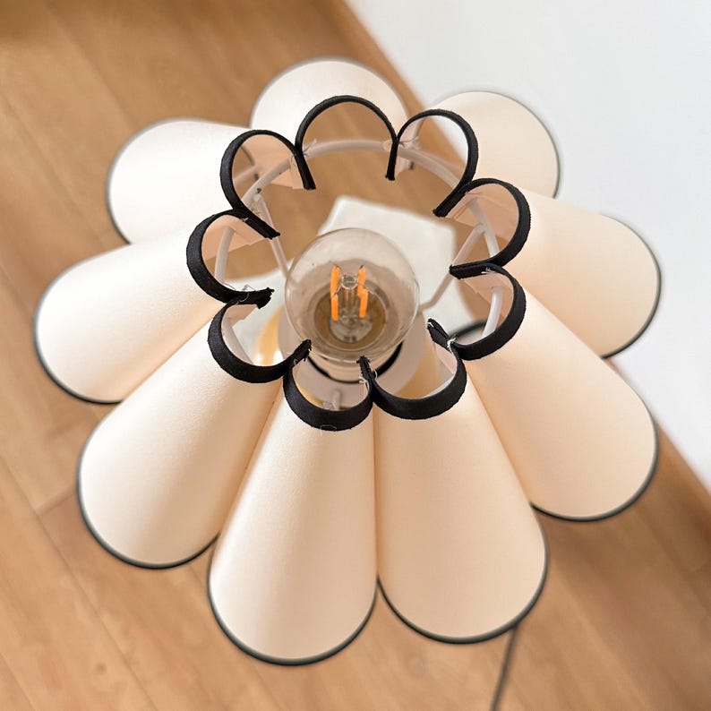 May include: A white lampshade with black trim, shaped like a flower with multiple petals. The lampshade is lit with a light bulb.