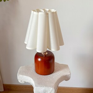 May include: A white lampshade with a pleated design sits atop a wooden base. The base is a rich brown color and has a cylindrical shape. The lamp is on a white pedestal.