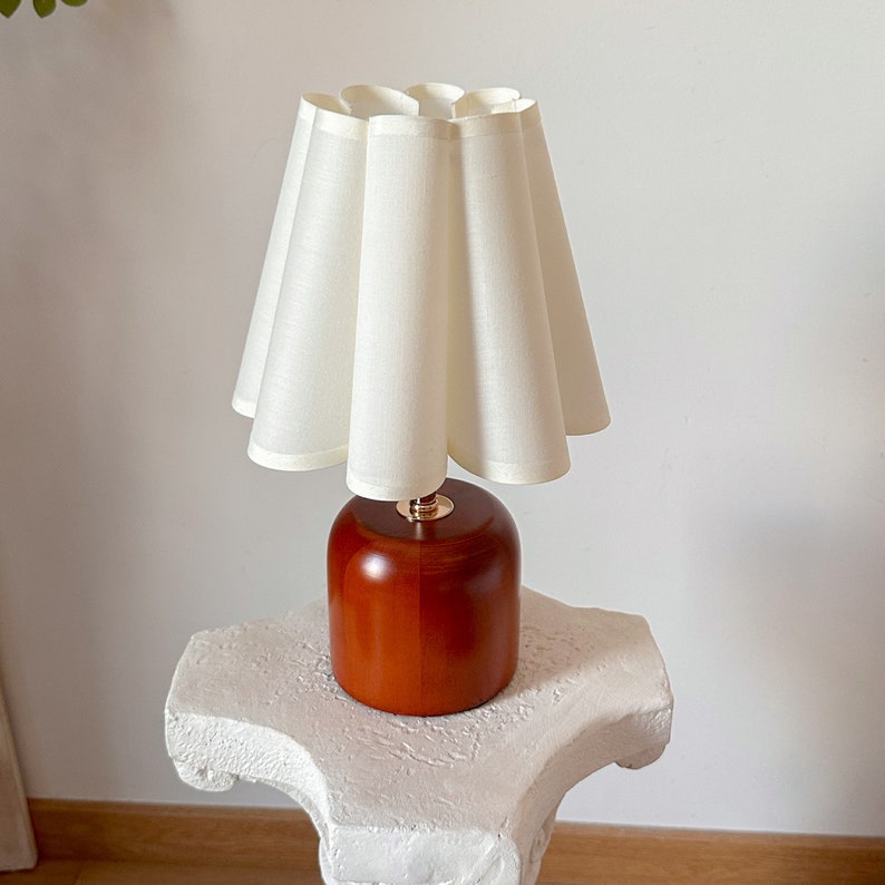 May include: A white lampshade with a pleated design sits atop a wooden base. The base is a rich brown color and has a cylindrical shape. The lamp is on a white pedestal.