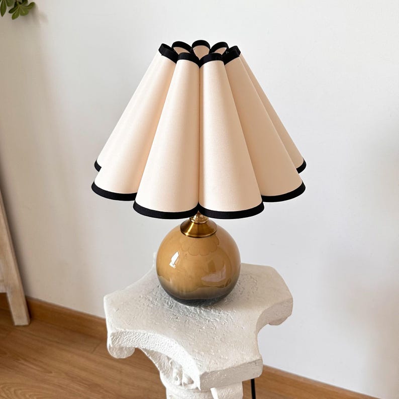 May include: A table lamp with a cream-colored pleated lampshade trimmed in black. The lamp base is a round, brown ceramic sphere.