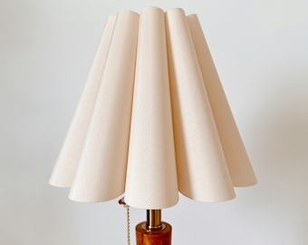 Puede incluir: Una lámpara de mesa con una pantalla de tela plisada de color crema y forma cónica. La base de la lámpara es de madera pulida con forma cilíndrica, acentuada por un cuello de metal dorado y un interruptor de cadena a juego.