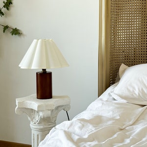May include: A table lamp featuring a cylindrical dark brown wooden base and a pleated off-white fabric lampshade. The lamp is positioned on a white stone pedestal with carved detailing, standing next to a bed with white bedding and a cane headboard.