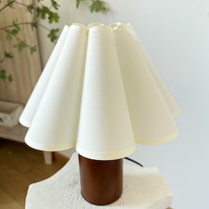 May include: A table lamp featuring a cylindrical brown base and a pleated, off-white fabric lampshade. The shade has a distinct scalloped edge design, and the lamp includes a black power cord extending from the base of the unit.