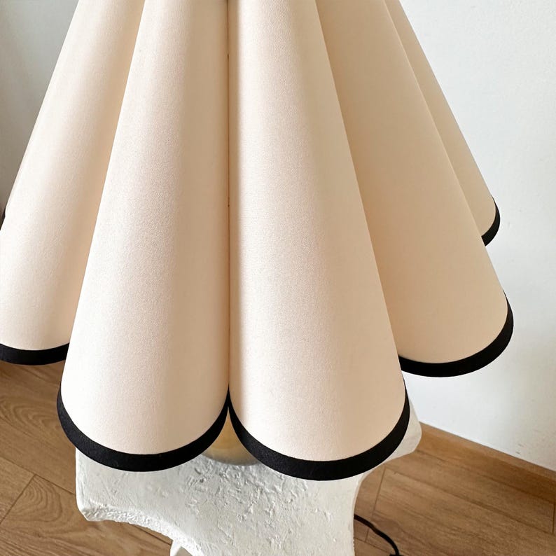 May include: A white lampshade with black trim, featuring a pleated design. The lampshade is attached to a white base.