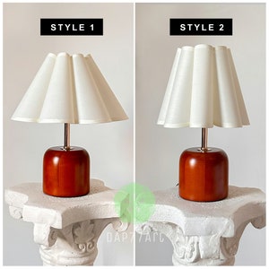 May include: Two table lamps with wooden bases and white fabric lampshades. The lampshades have a pleated design. The lamp on the left has a more rounded shade, while the lamp on the right has a more pointed shade. The text "STYLE 1" is above the lamp on the left, and the text "STYLE 2" is above the lamp on the right.