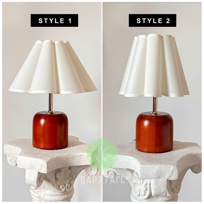 May include: Two table lamps with wooden bases and white fabric lampshades. The lampshades have a pleated design. The lamp on the left has a more rounded shade, while the lamp on the right has a more pointed shade. The text "STYLE 1" is above the lamp on the left, and the text "STYLE 2" is above the lamp on the right.