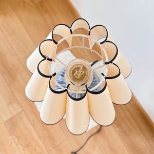 May include: A cream-colored table lamp with a floral-shaped lampshade. The lampshade has a white metal frame and is trimmed with black edging. The lamp sits on a white surface and has a visible lightbulb.