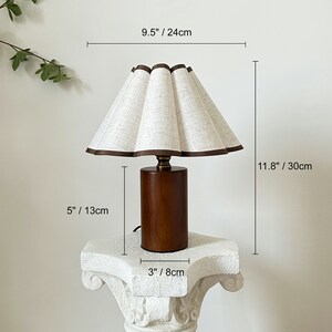 K&ouml;nnte beinhalten: Eine Tischlampe aus Holz mit einem wei&szlig;en Leinenlampenschirm. Der Lampenschirm hat einen gewellten Rand und einen braunen Besatz. Der Lampenfu&szlig; ist ein dunkelbrauner Zylinder mit einem goldfarbenen Abschluss. Die Lampe ist 30 cm hoch und der Lampenschirm 24 cm breit.