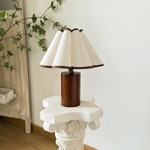 K&ouml;nnte beinhalten: Eine Tischlampe aus Holz mit einem wei&szlig;en Leinenlampenschirm. Der Lampenschirm hat einen braunen Besatz und ist gefaltet. Die Lampe steht auf einem wei&szlig;en Sockel.