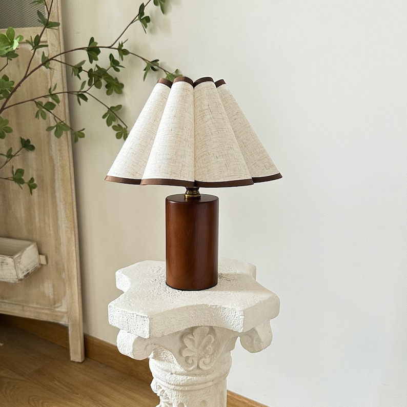 K&ouml;nnte beinhalten: Eine Tischlampe aus Holz mit einem wei&szlig;en Leinenlampenschirm. Der Lampenschirm hat einen braunen Besatz und ist gefaltet. Die Lampe steht auf einem wei&szlig;en Sockel.