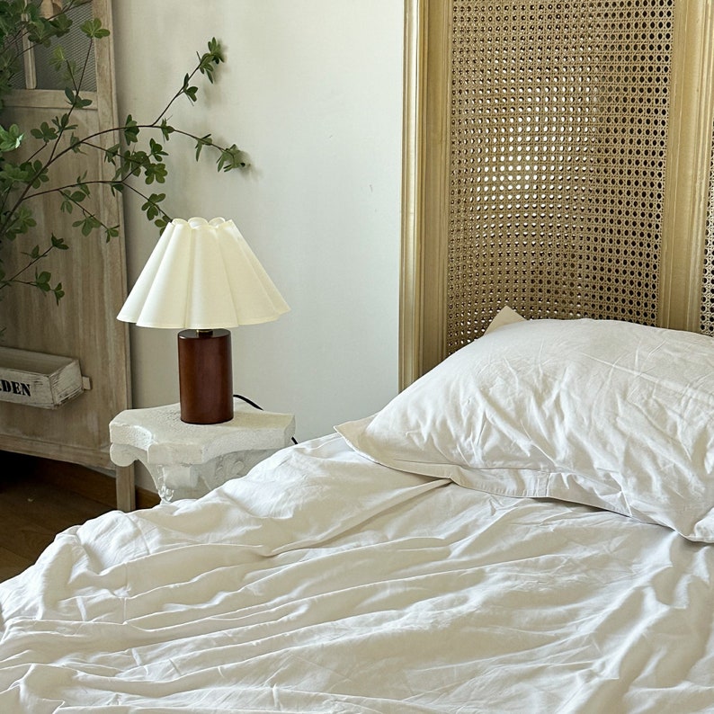 May include: A table lamp with a cylindrical dark brown wooden base and a pleated off-white fabric lampshade. The lamp sits on a white stone pedestal next to a bed with white bedding and a headboard featuring a woven cane panel design.