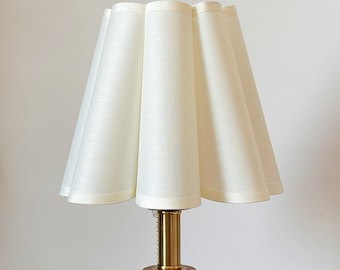 Puede incluir: Una lámpara de mesa con una pantalla de tela plisada en color blanco roto de textura lisa. La base de la lámpara consta de un cuello cilíndrico de latón pulido conectado a un soporte cilíndrico de madera con veta natural visible.