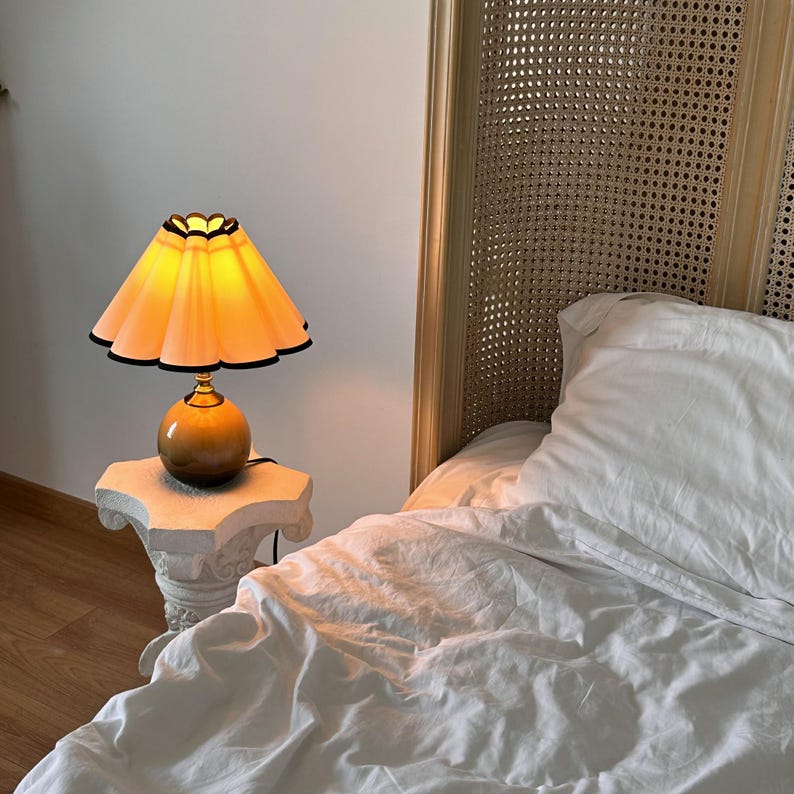May include: A bedside table lamp with a white base and a ruffled, peach-colored lampshade with a black trim. The lamp is turned on and illuminating the white bedding of a bed with a woven headboard.
