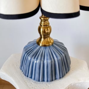May include: A decorative table lamp featuring a light blue, ribbed ceramic base and a brass stem. The lamp shade is cream-colored with a black trim. The lamp is a stylish accent piece for any room.