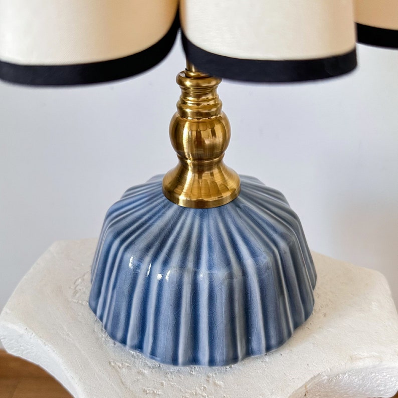 May include: A decorative table lamp featuring a light blue, ribbed ceramic base and a brass stem. The lamp shade is cream-colored with a black trim. The lamp is a stylish accent piece for any room.
