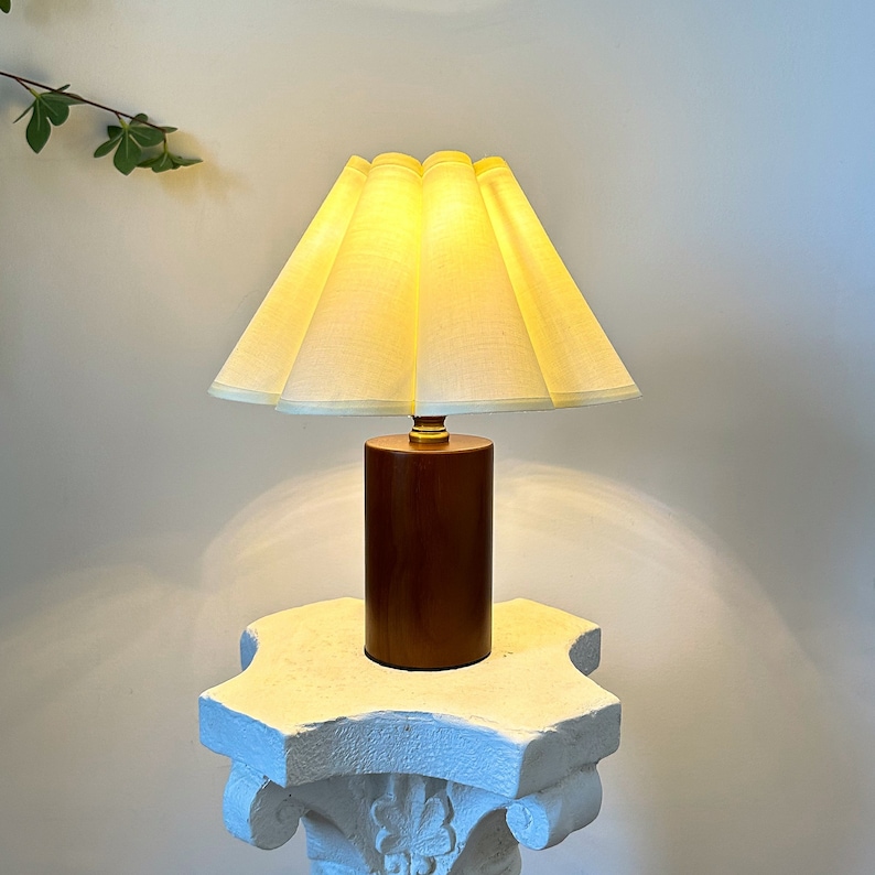 May include: A table lamp featuring a cylindrical dark brown wooden base and a pleated, light yellow fabric lampshade. The lamp emits a warm, soft glow from beneath the shade, which is attached to the base by a small, metallic gold-colored connector.