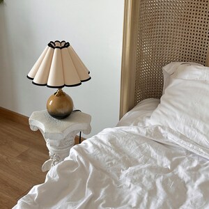 May include: A bedside table lamp with a white pleated lampshade with a black trim. The lamp has a brown and gold ceramic base. The lamp is sitting on a white pedestal table.