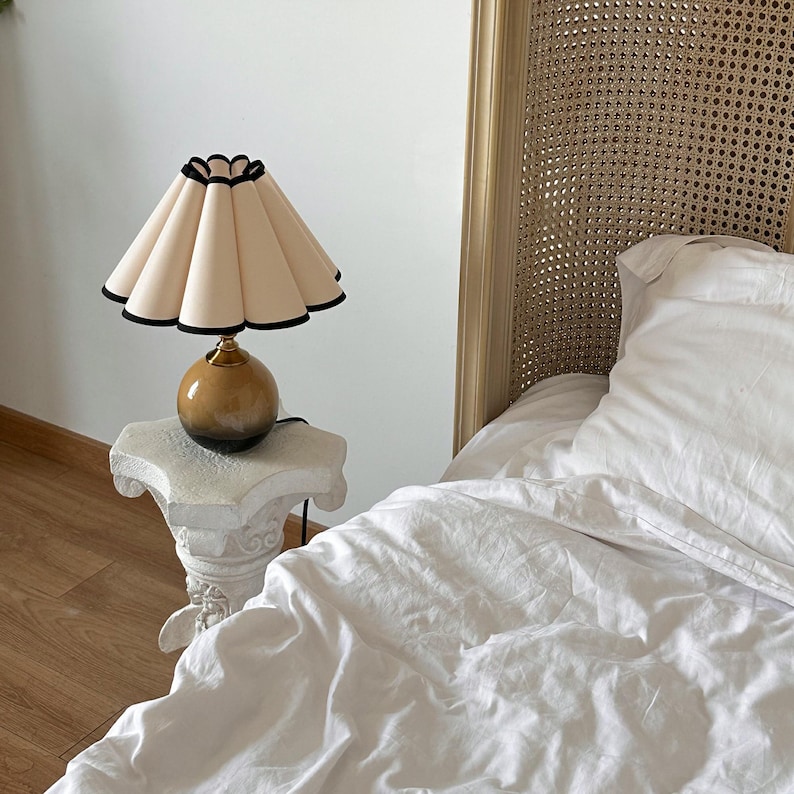 May include: A bedside table lamp with a white pleated lampshade with a black trim. The lamp has a brown and gold ceramic base. The lamp is sitting on a white pedestal table.