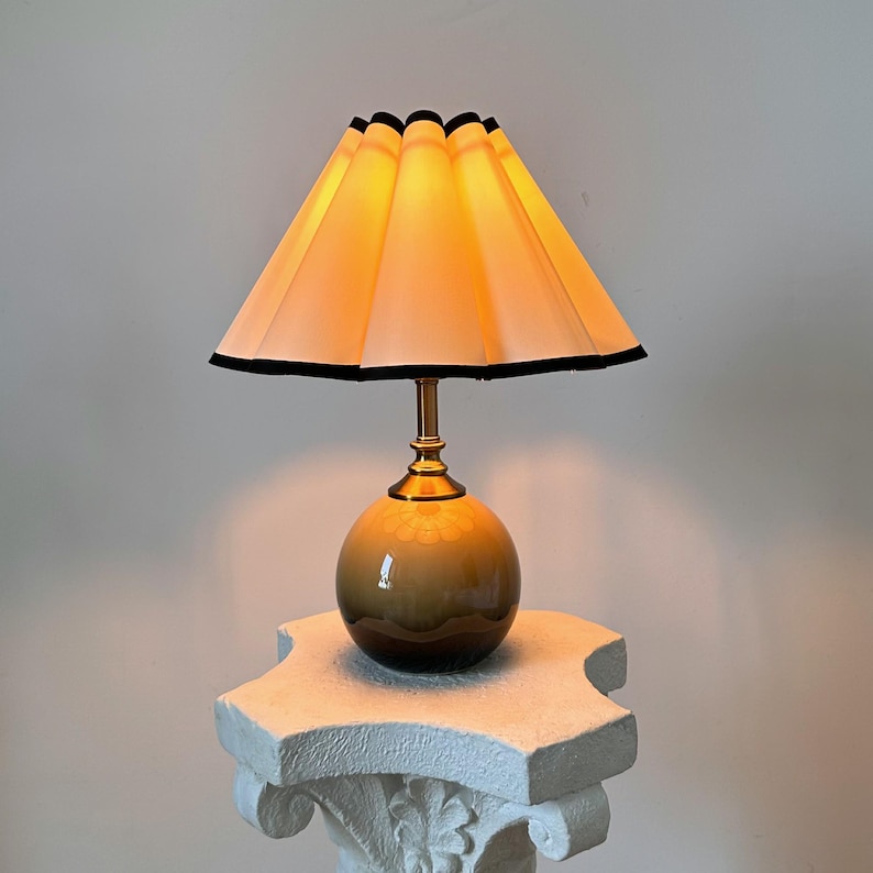 May include: A table lamp with a pleated, off-white lampshade with a black trim. The lamp base is a round, brown ceramic sphere.
