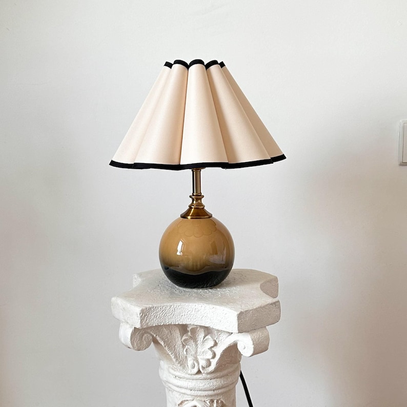 May include: A table lamp with a white pleated lampshade with a black trim and a brown and black ceramic base.