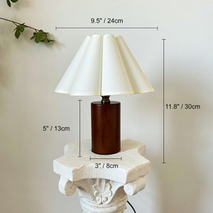 May include: A table lamp with a cylindrical dark wood base and a pleated off-white fabric shade. The lamp measures 11.8 inches in total height and 9.5 inches in shade width. The base is 5 inches tall and 3 inches wide, featuring a brass-toned neck.