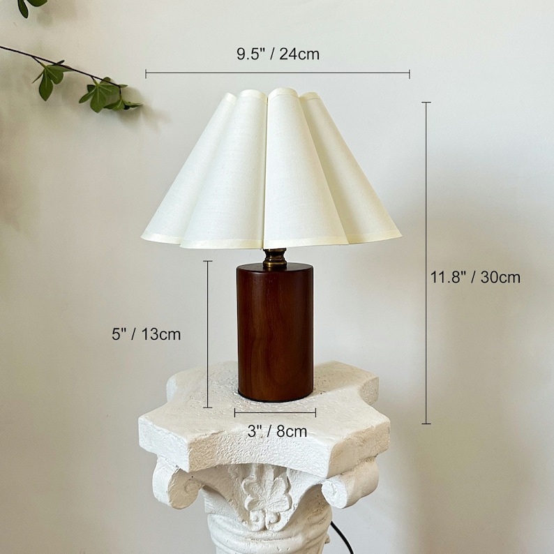 May include: A table lamp with a cylindrical dark wood base and a pleated off-white fabric shade. The lamp measures 11.8 inches in total height and 9.5 inches in shade width. The base is 5 inches tall and 3 inches wide, featuring a brass-toned neck.