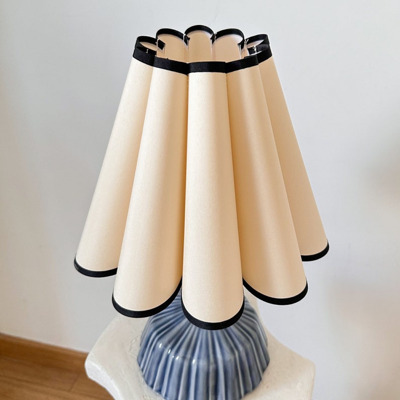 May include: A decorative table lamp with a cream-colored shade featuring a scalloped design and black trim. The lamp sits on a blue ceramic base with a ribbed texture. The lamp is a stylish home decor item.