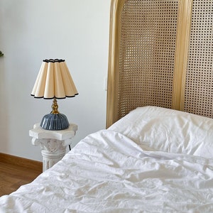 May include: A cream-colored pleated lampshade with a black trim sits atop a blue ceramic base with gold accents. The lamp is placed on a white, ornate pedestal. A woven headboard and white bedding are in the background.