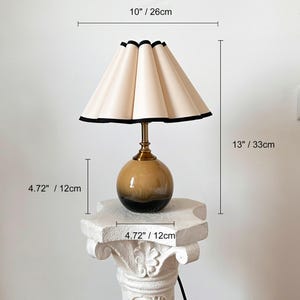 May include: A table lamp with a cream-colored pleated lampshade with a black trim. The lamp base is a round, brown and black ceramic globe. The lamp is 13 inches tall and the base is 4.72 inches in diameter.