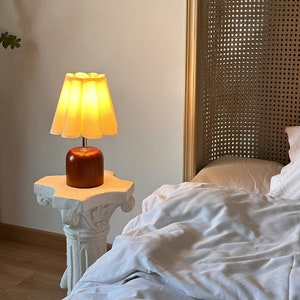 May include: A bedside table lamp with a white pleated lampshade and a brown wooden base. The lamp is turned on and illuminating the room.