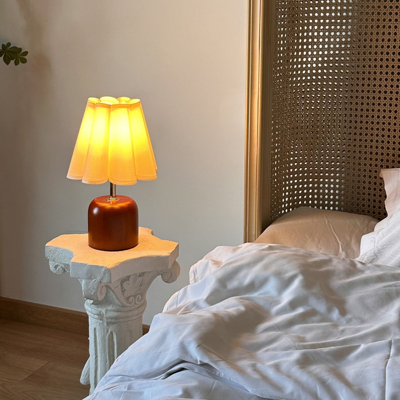 May include: A bedside table lamp with a white pleated lampshade and a brown wooden base. The lamp is turned on and illuminating the room.