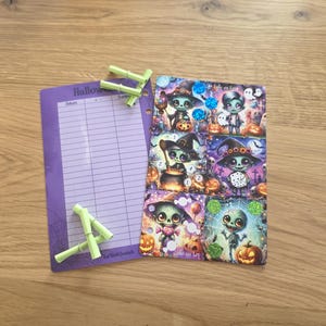 May include: A purple Halloween-themed notepad with lined pages and a decorative spiderweb design, next to a sheet of Halloween-themed stickers featuring cartoon characters and pumpkins. Several green bamboo sticks are also present.