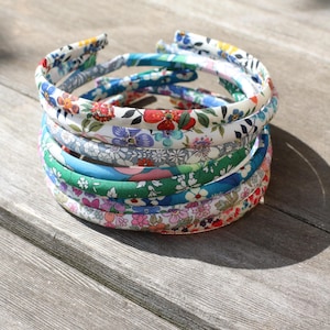 May include: A stack of six thin fabric-covered headbands featuring various floral patterns in colors including red, blue, green, pink, and white. Each headband has a rounded profile and is wrapped in a smooth textile with intricate botanical prints.