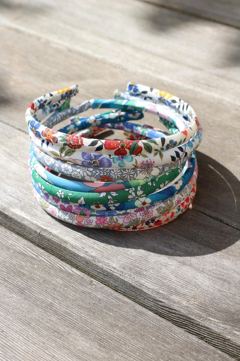 May include: A stack of six thin fabric-covered headbands featuring various floral patterns in colors including red, blue, green, pink, and white. Each headband has a rounded profile and is wrapped in a smooth textile with intricate botanical prints.