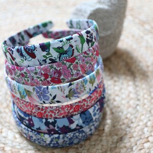 May include: A stack of six fabric-covered headbands featuring various floral and botanical patterns in colors including blue, pink, red, green, and white. The headbands are arranged vertically, showcasing distinct textile designs on each individual piece.