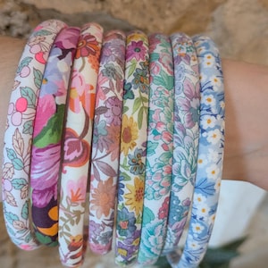 May include: A collection of eight thin, fabric-covered headbands arranged in a row. Each headband features a distinct floral pattern with various color palettes including pink, orange, yellow, green, purple, and blue, showcasing intricate botanical prints.
