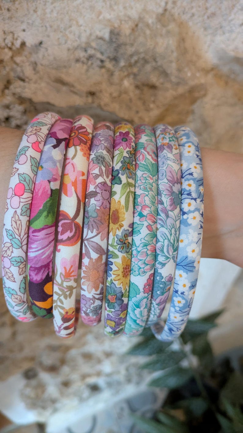May include: A collection of eight thin, fabric-covered headbands arranged in a row. Each headband features a distinct floral pattern with various color palettes including pink, orange, yellow, green, purple, and blue, showcasing intricate botanical prints.