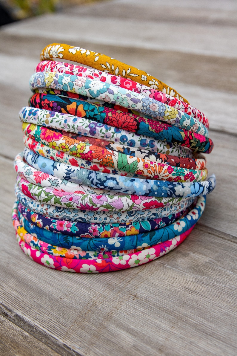May include: A stack of twelve fabric-covered headbands featuring various floral patterns in colors including mustard yellow, pink, blue, red, and white. Each headband has a rounded profile and is wrapped in cotton fabric with intricate botanical prints.