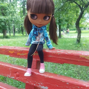 Puede incluir: Una mu&ntilde;eca con el pelo largo y casta&ntilde;o que lleva una sudadera con capucha azul y negra con estampado de galaxia, leggings negros y zapatillas rosas. La mu&ntilde;eca est&aacute; sentada en un banco de madera rojo.