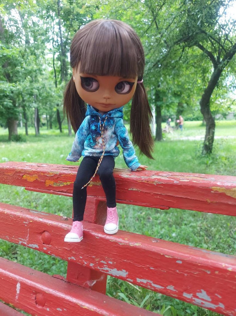 Puede incluir: Una mu&ntilde;eca con el pelo largo y casta&ntilde;o que lleva una sudadera con capucha azul y negra con estampado de galaxia, leggings negros y zapatillas rosas. La mu&ntilde;eca est&aacute; sentada en un banco de madera rojo.