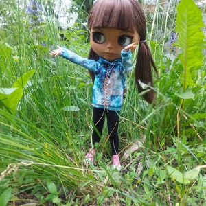 Puede incluir: Una mu&ntilde;eca con cabello casta&ntilde;o y ojos azules que lleva una sudadera con capucha de estampado de galaxia azul y marr&oacute;n y pantalones negros. La mu&ntilde;eca est&aacute; de pie en un campo de hierba verde.