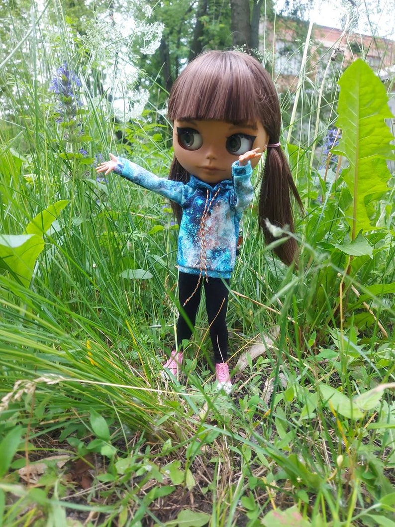 Puede incluir: Una mu&ntilde;eca con cabello casta&ntilde;o y ojos azules que lleva una sudadera con capucha de estampado de galaxia azul y marr&oacute;n y pantalones negros. La mu&ntilde;eca est&aacute; de pie en un campo de hierba verde.