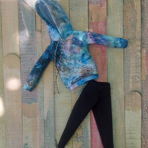 Puede incluir: Una sudadera con capucha azul y gris con estampado de galaxia y leggings negros. La ropa est&aacute; colocada sobre una superficie de madera.