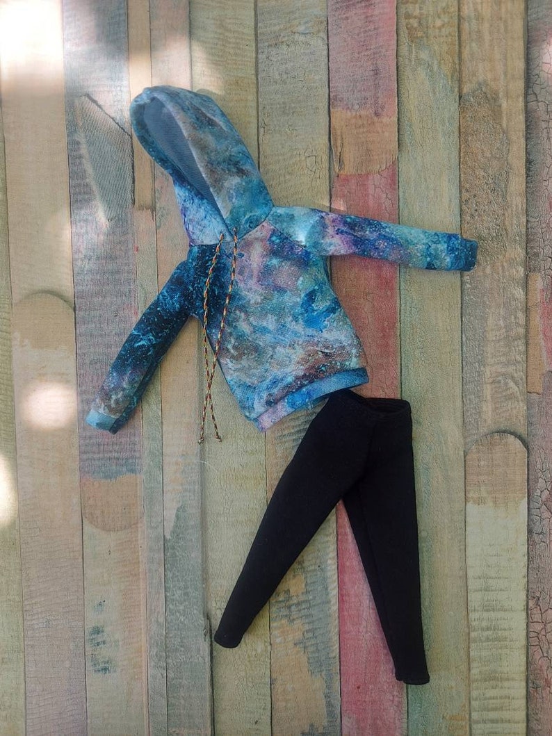 Puede incluir: Una sudadera con capucha azul y gris con estampado de galaxia y leggings negros. La ropa est&aacute; colocada sobre una superficie de madera.