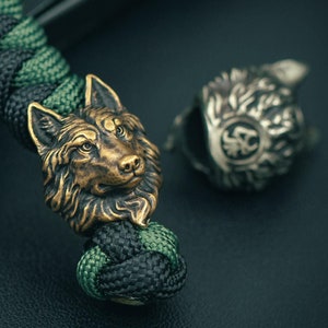 May include: A black and green paracord bracelet with a bronze wolf head charm. The charm is detailed and realistic, with a textured surface.